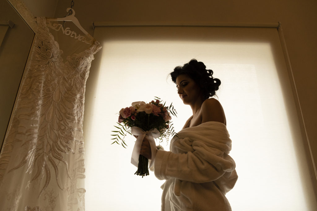 mulher segurando seu buquê de noiva em frente ao vestido pendurado.